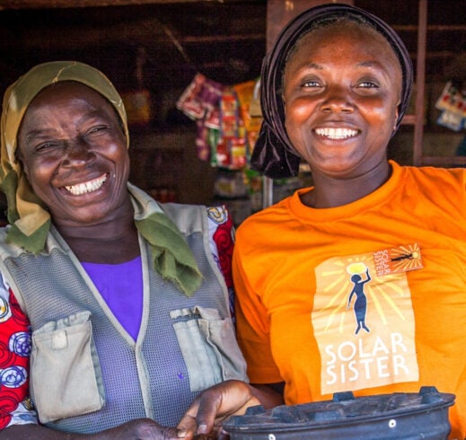Two women smile outside a small shop; the woman on the right wears a yellow Solar Sister T-shirt and holds a black cooking pot, while the woman on the left stands beside her in a headscarf and patterned blouse.
