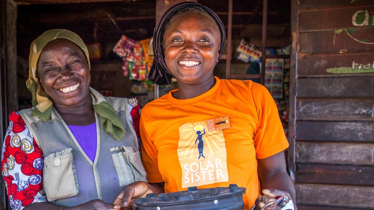 Two women smile outside a small shop; the woman on the right wears a yellow Solar Sister T-shirt and holds a black cooking pot, while the woman on the left stands beside her in a headscarf and patterned blouse.